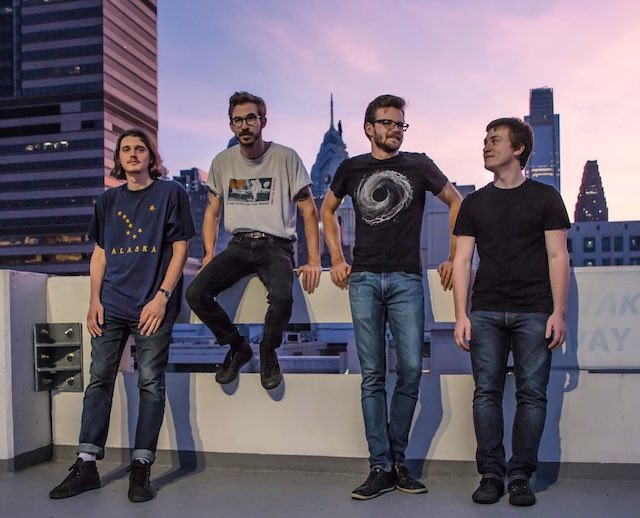 The band sitting along a parking garage rooftop with the Philadelphia skyline behind them.